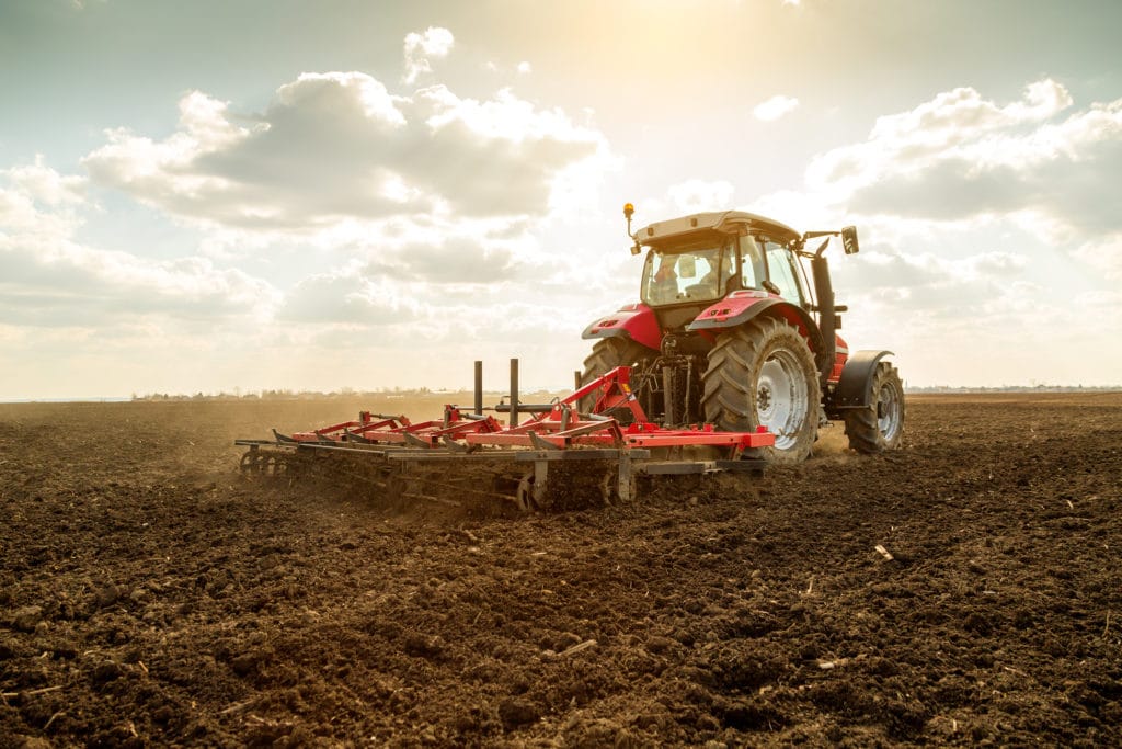 A tractor pulling a seedbed cultivator across a bare field under a bright sky
